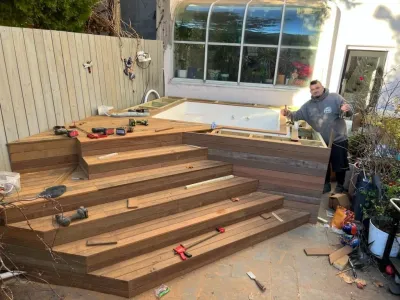 A worker gives a thumbs-up next to a partially constructed multi-level wooden deck surrounding a new white hot tub in a backyard setting.