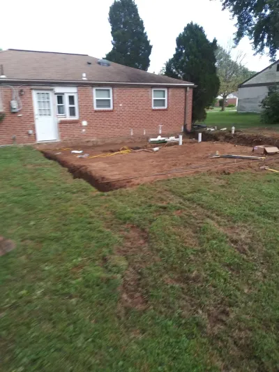 A residential backyard shows an excavated area next to a brick house, revealing utility pipes, electrical cables, and rebar for a new foundation.
