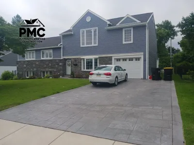A completed residential exterior featuring blue horizontal siding, dark stone veneer, white trim, and a newly installed stamped concrete driveway.