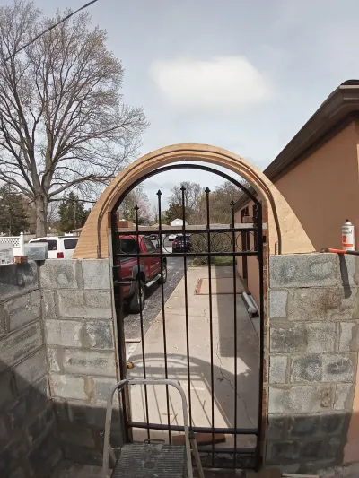 An arched entryway is under construction, featuring unfinished concrete block walls, a black metal gate, and wooden formwork for the arch overhead, with construction tools visible nearby.