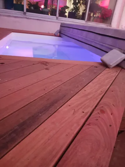 A built-in hot tub with blue and purple underwater lighting is surrounded by reddish-brown wooden decking, partially covered by a gray spa cover.