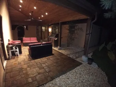 A covered outdoor patio features paver flooring, comfortable seating with red cushions, a ceiling fan, and a built-in grill area, illuminated by warm recessed lighting at night.
