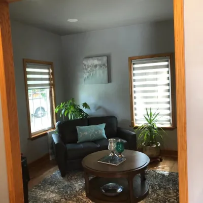 A cozy sitting area features a dark loveseat, a round coffee table, and two windows fitted with modern striped zebra blinds providing filtered light.