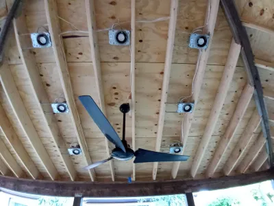 A view looking up at an exposed wood ceiling structure featuring a large black ceiling fan and multiple installed light fixtures with visible wiring.