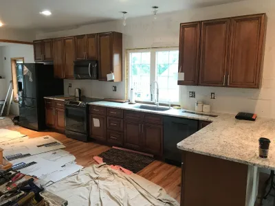 A residential kitchen renovation is underway, featuring newly installed dark wood cabinets, light granite countertops, and appliances, with construction materials visible on the wood floor.